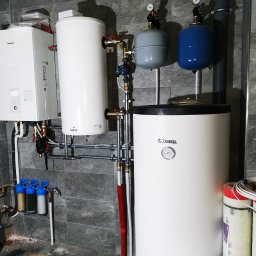 KOVALINSTAL JACEK KOWALIK - Kotłownia w szarościach: białe zbiorniki, filtry wody z niebieskimi obudowami, rury i zawory. Sprzęt zamontowany na tle szarej ściany imitującej kamień.