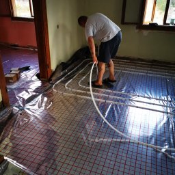 KOVALINSTAL JACEK KOWALIK - Instalacja ogrzewania podłogowego: mężczyzna układa białe rurki na folii z czerwoną siatką w surowym wnętrzu. Widoczne okna i drzwi do innego pomieszczenia.