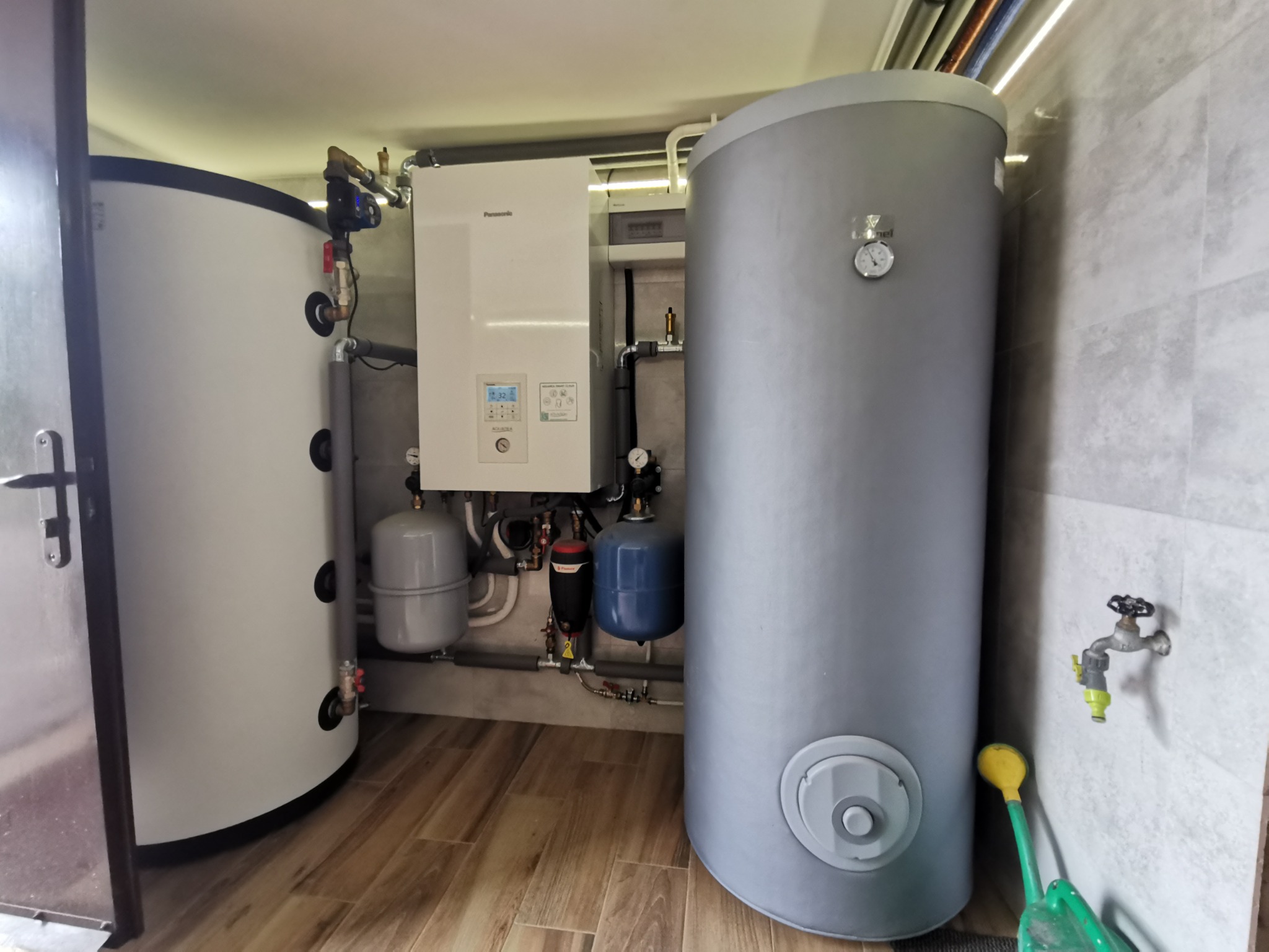 Zestaw urządzeń grzewczych: biały piec gazowy Panasonic z wyświetlaczem, dwa zbiorniki (biały i szary), naczynia przeponowe, instalacja hydrauliczna. Widok w pomieszczeniu z płytkami.