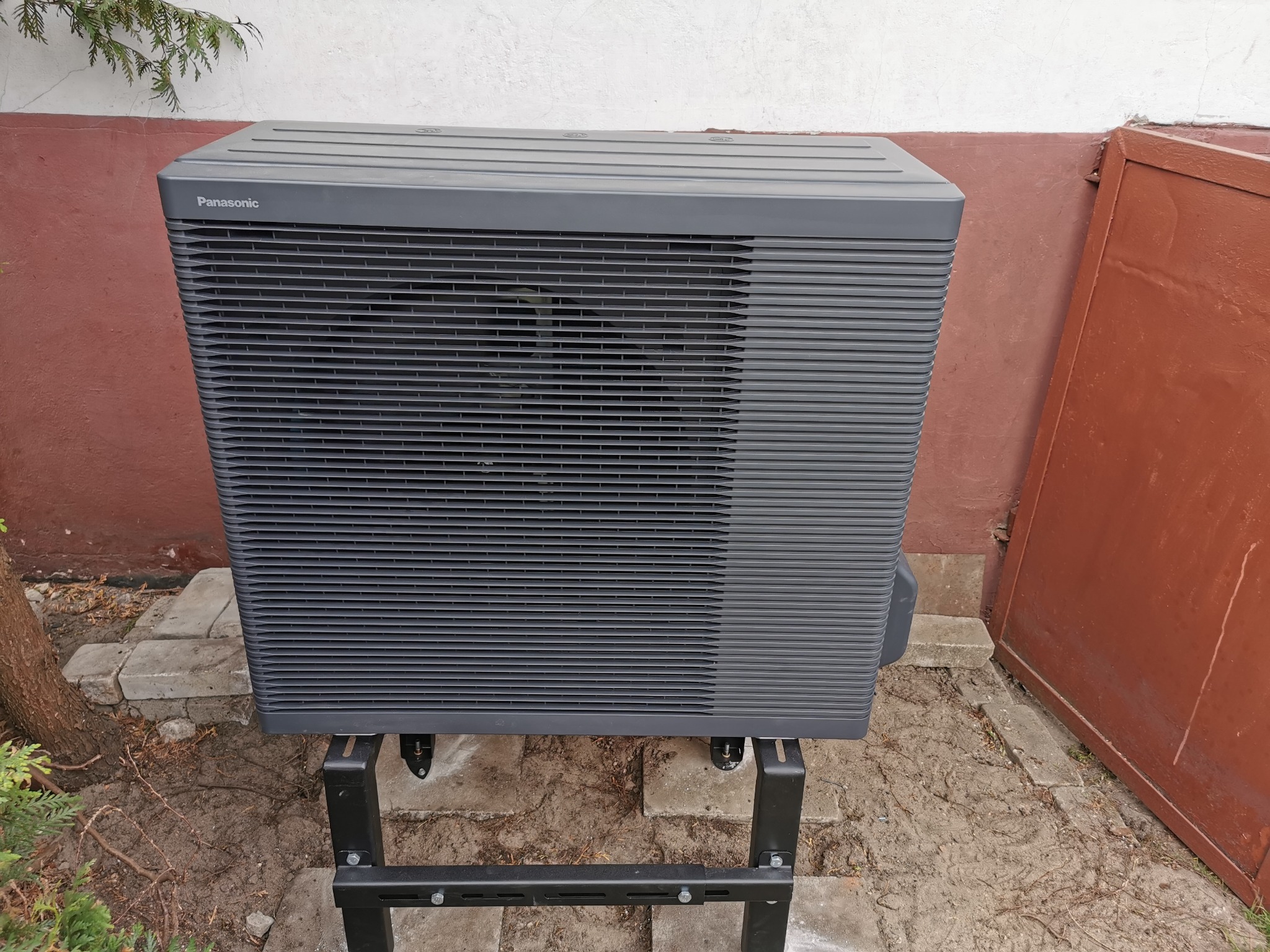 Zewnętrzna jednostka klimatyzacji Panasonic, ustawiona na czarnej metalowej podstawie, na tle ściany budynku z elementami zieleni. Widok z bliska, skupiony na urządzeniu.