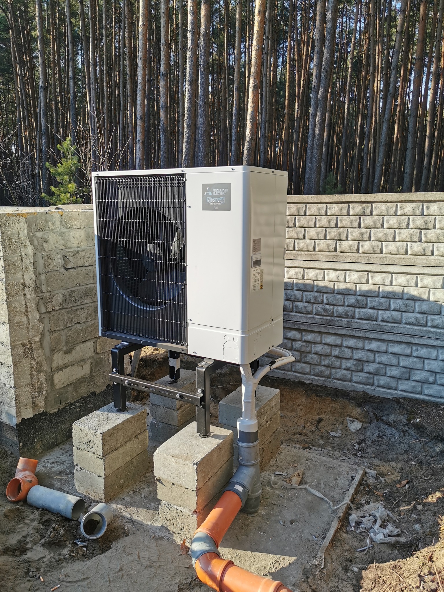 Biała pompa ciepła Mitsubishi Electric na zewnątrz, zamontowana na czarnej metalowej konstrukcji, z widocznymi rurami. W tle las i betonowy murek.