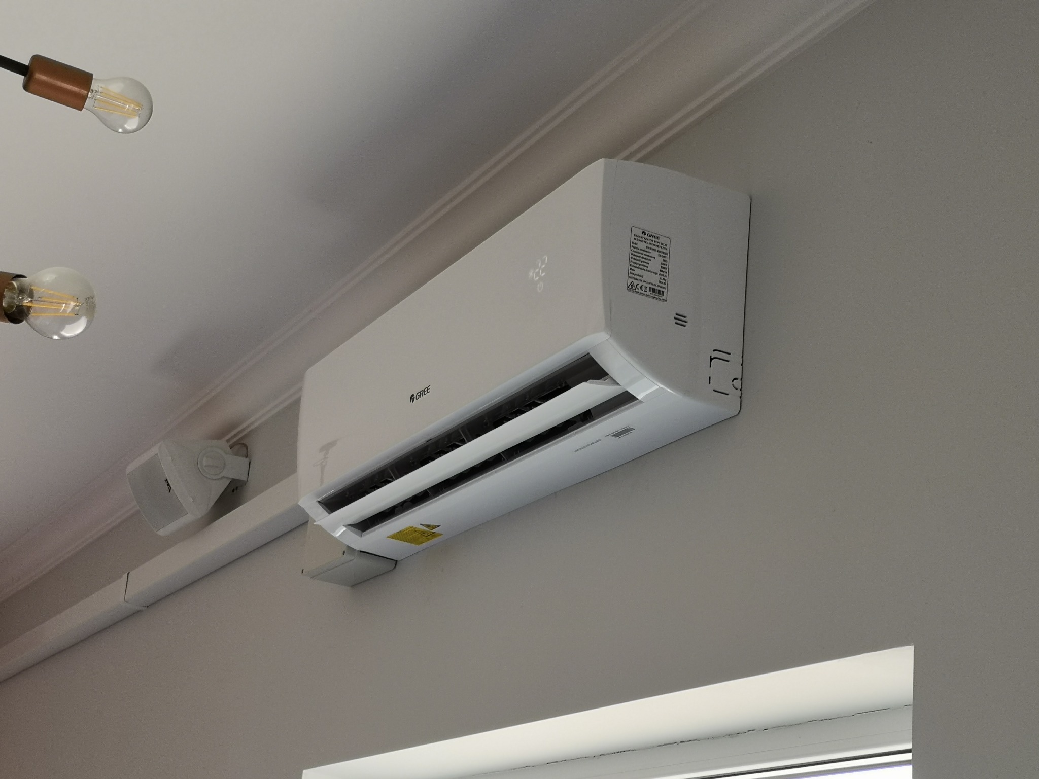 Biała klimatyzacja Gree zamontowana na szarej ścianie w pomieszczeniu. Widoczny fragment okna i lampy w stylu loft.