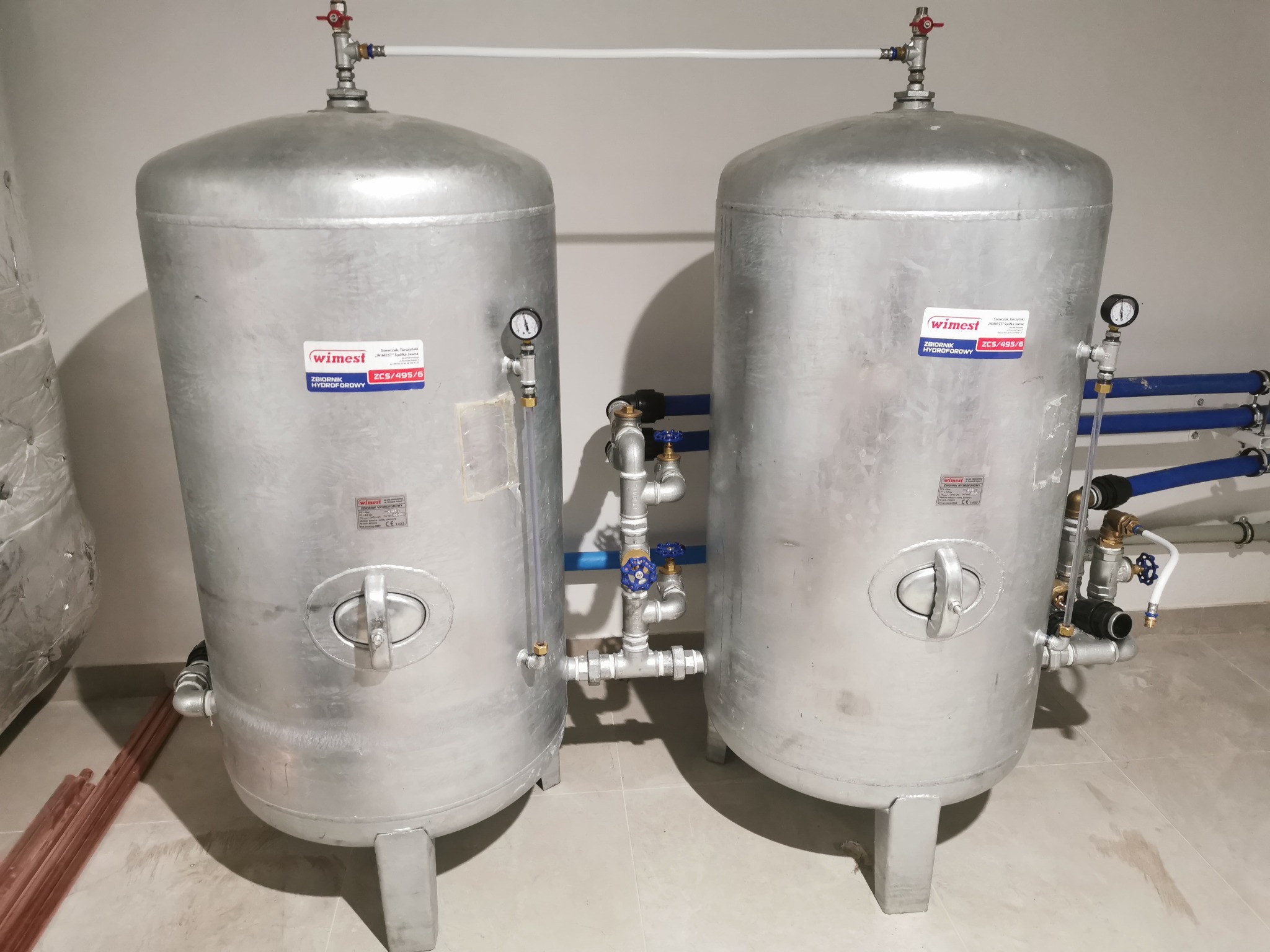 Dwa srebrne, pionowe zbiorniki hydroforowe WIMEST ZCS/495/6 z manometrami i rurami, z bliska. Widoczne rury miedziane obok zbiornika i biała ściana w tle.