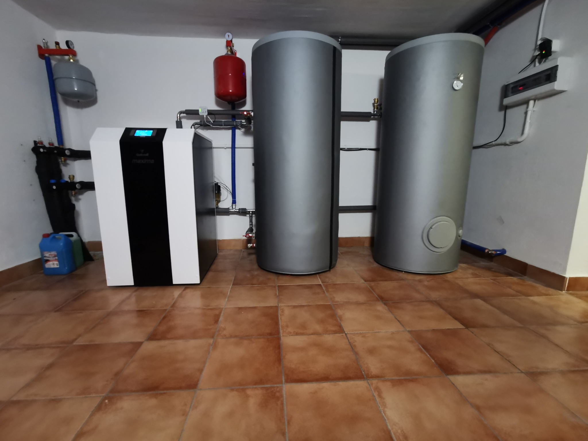 Instalacja grzewcza w kotłowni: nowoczesny piec z wyświetlaczem, dwa szare zbiorniki akumulacyjne, instalacja rurowa z czerwonymi zbiornikami wyrównawczymi.