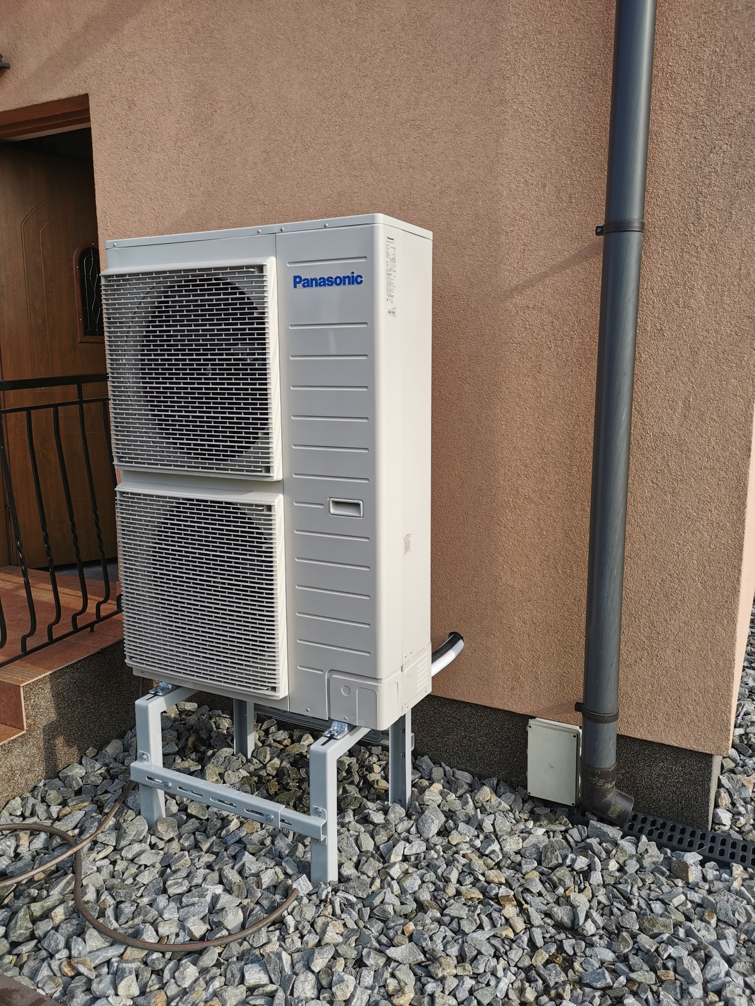Biała, dwuczęściowa pompa ciepła Panasonic na metalowej konstrukcji, zamontowana przy ścianie budynku z rurą spustową. Podłoże wyłożone kamieniami.