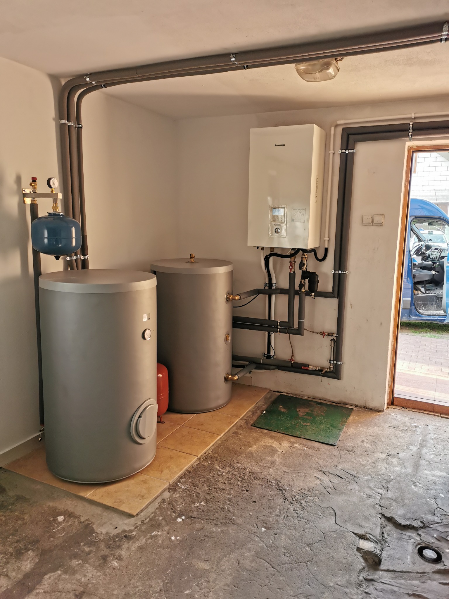 Kompletna instalacja CO z dwoma zbiornikami akumulacyjnymi, piecem gazowym Panasonic i orurowaniem w piwnicy. Widoczny otwarty samochód dostawczy.