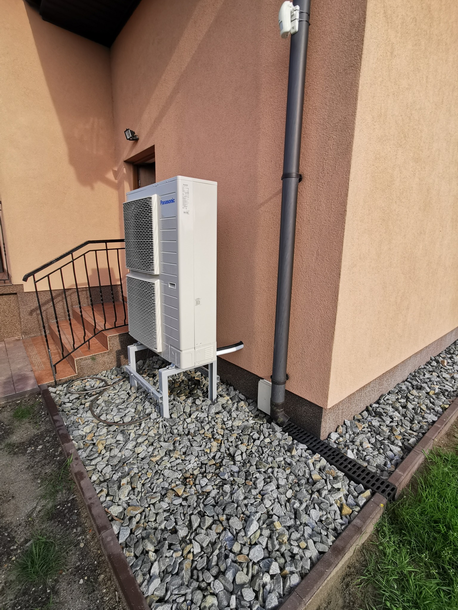 Biała, dwuczęściowa pompa ciepła Panasonic, zamontowana na metalowym stelażu przy ścianie budynku z brązową rurą spustową i kamiennym podłożem.