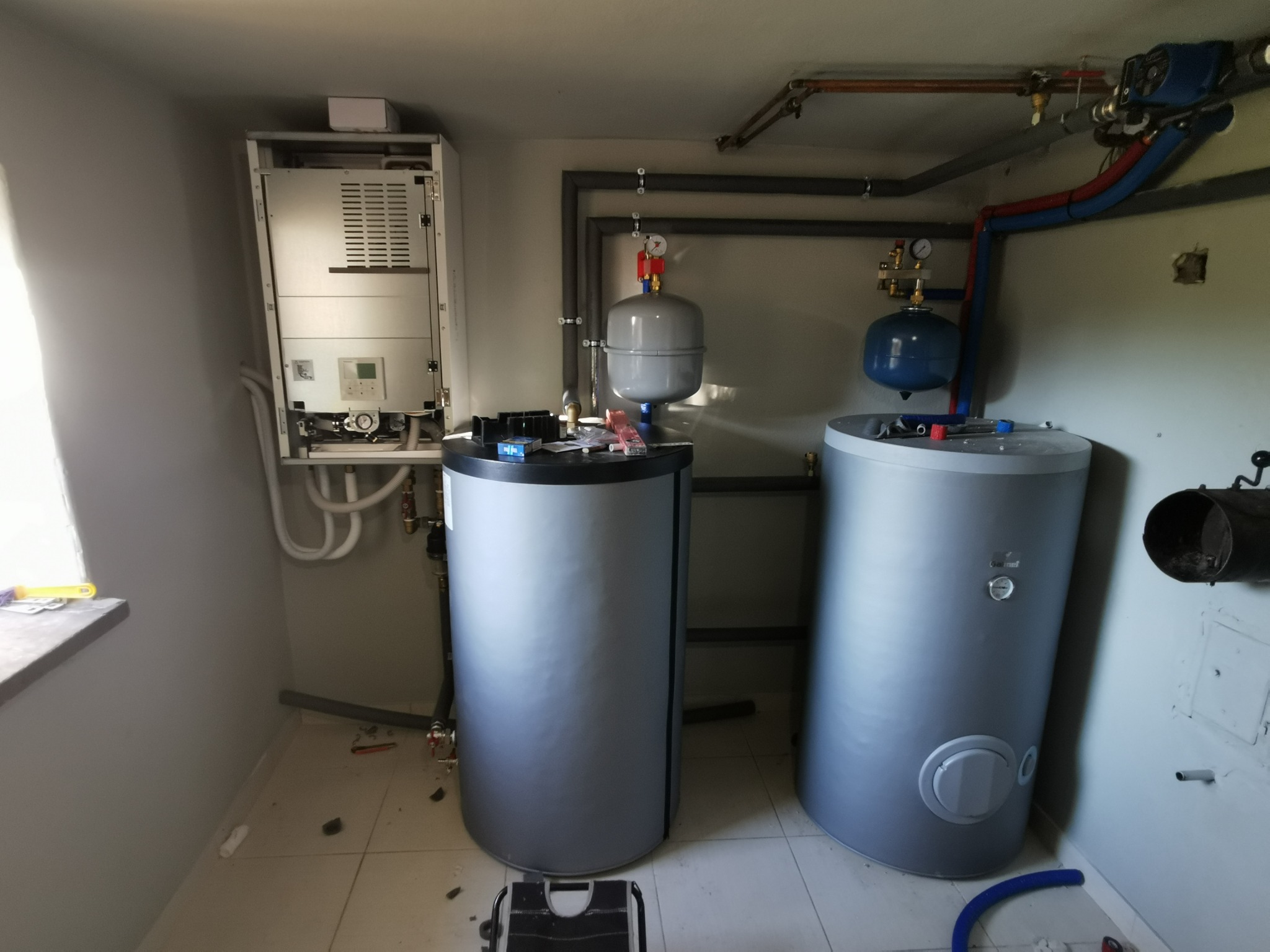 Zestaw grzewczy z dwoma zbiornikami akumulacyjnymi, kotłem i orurowaniem w jasnym pomieszczeniu. Widoczne manometry i zawory.