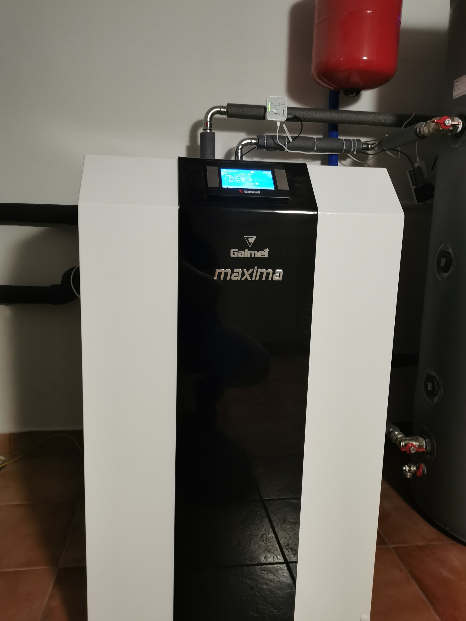 Biała pompa ciepła Galmet Maxima z czarnym panelem, widoczny zbiornik buforowy i instalacja rurowa w pomieszczeniu z brązowymi płytkami podłogowymi.