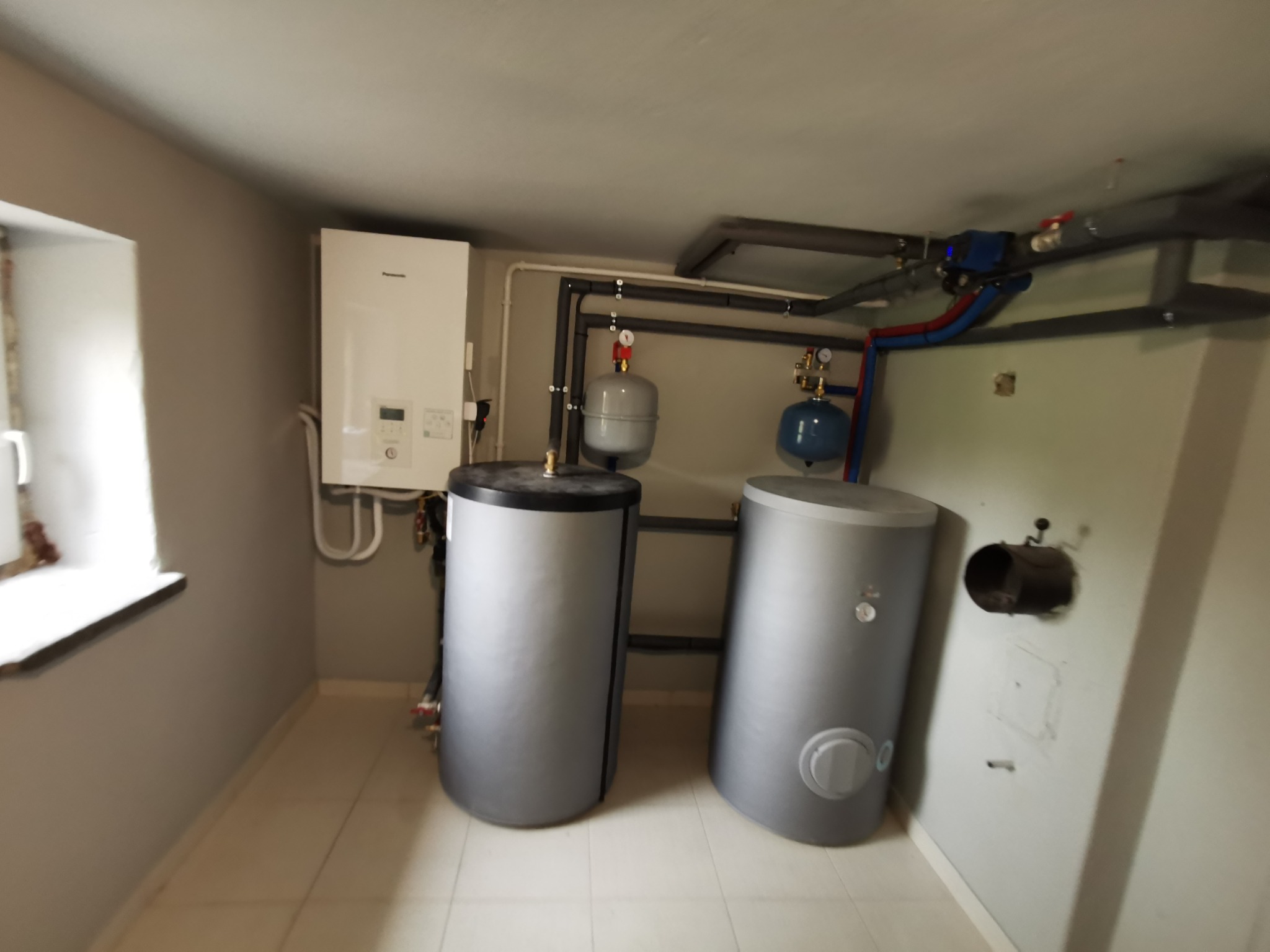 Kotłownia z dwoma szarymi zbiornikami akumulacyjnymi, białym piecem gazowym i instalacją rurową. Widok na okno i białe płytki podłogowe.