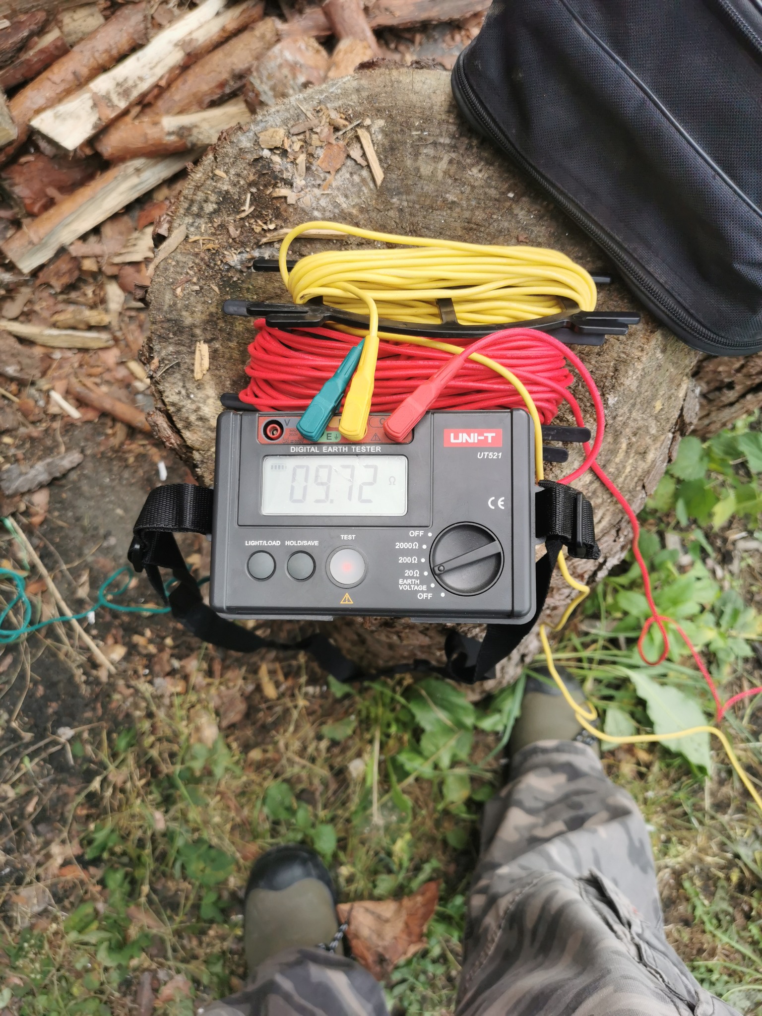 Miernik rezystancji uziemienia UNI-T UT521 na pniu, z rozłożonymi kablami pomiarowymi (czerwony, żółty), obok pokrowiec. Widoczne fragmenty butów osoby wykonującej pomiar.