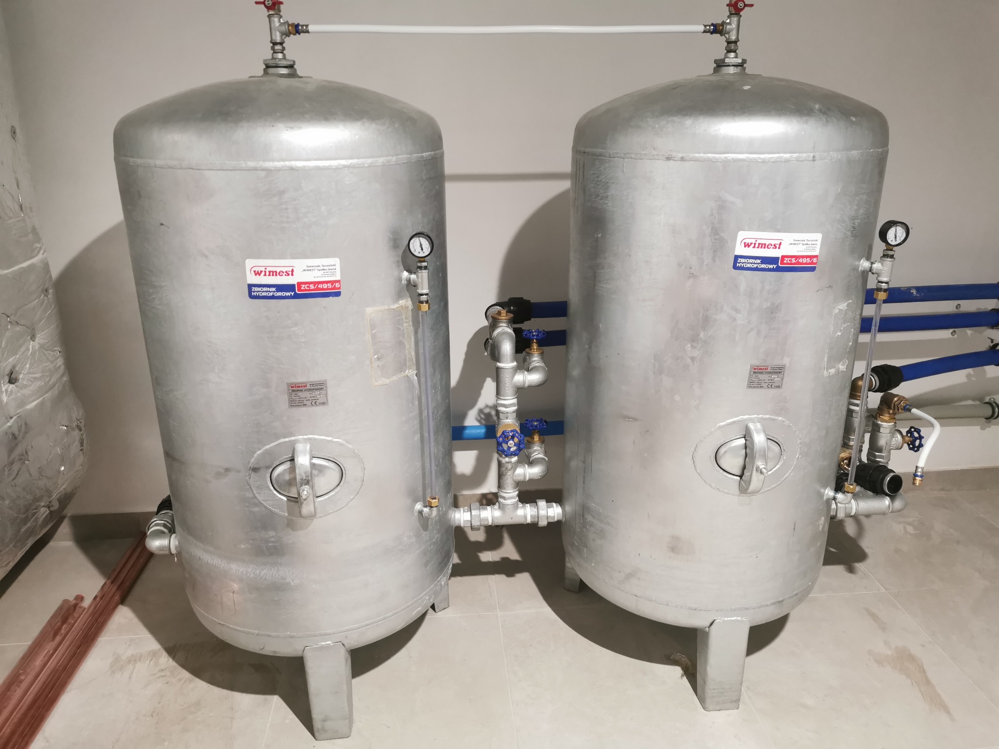 Dwa srebrne zbiorniki hydroforowe WIMEST ZCS/495/6 z manometrami i instalacją hydrauliczną na jasnej podłodze, w pomieszczeniu z białymi ścianami.