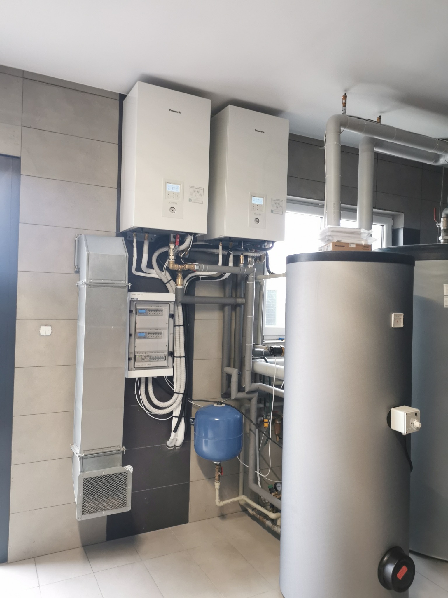 Dwie białe pompy ciepła Panasonic zamontowane na ścianie z szarą płytką. Widoczna instalacja, zbiornik wody, skrzynka elektryczna i kanał wentylacyjny.