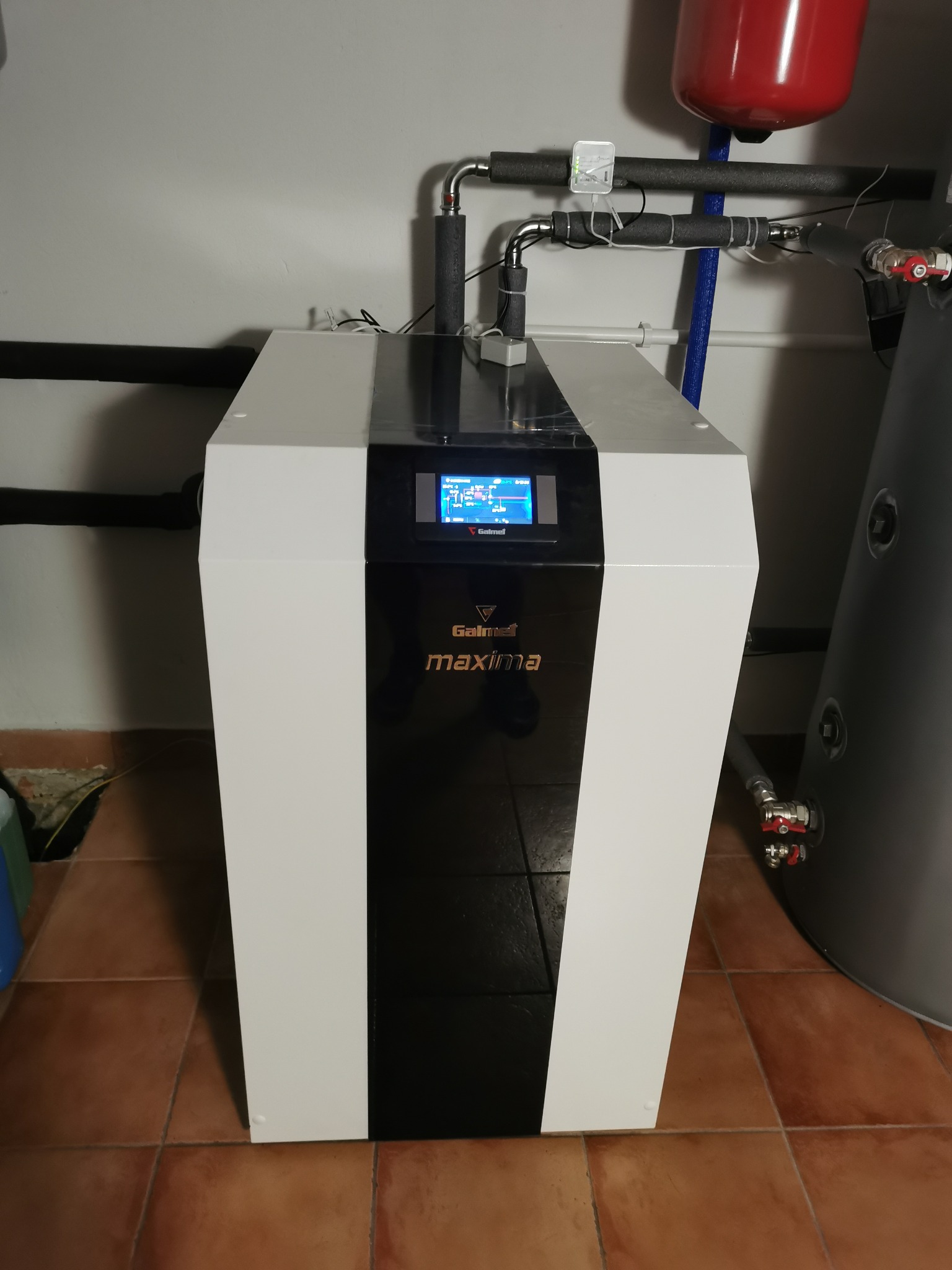 Biało-czarna pompa ciepła Galmet Maxima z ekranem, widoczne rury instalacji i zbiornik. Ujęcie w kotłowni na brązowych płytkach.