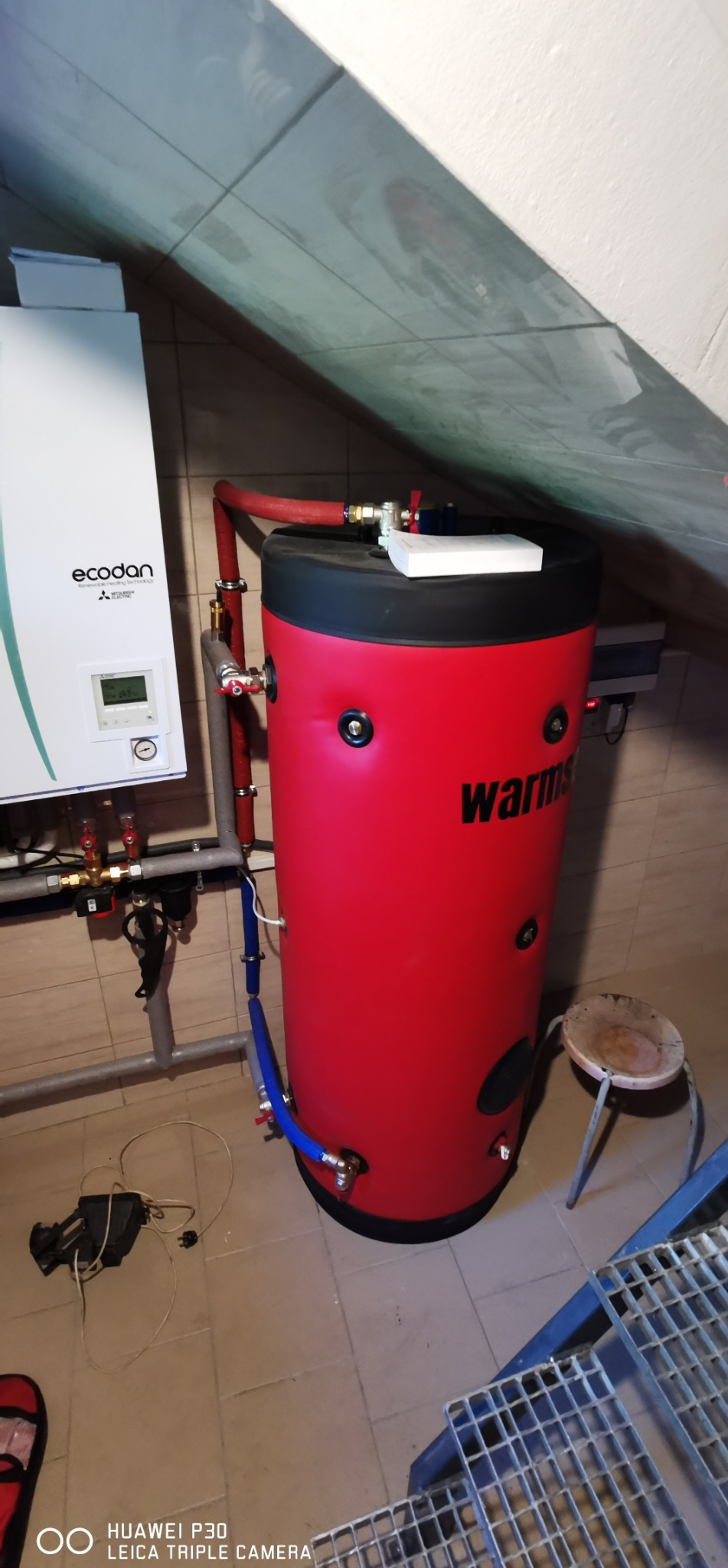 Czerwony zasobnik ciepłej wody użytkowej 'Warms' w kotłowni na poddaszu, obok jednostki zewnętrznej pompy ciepła Mitsubishi Ecodan i krzesła. Widoczne rury instalacji.