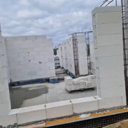 Wnętrze surowego budynku z betonu komórkowego w trakcie budowy. Widoczne ściany, zbrojenia, wózek z bloczkami i mokra posadzka.