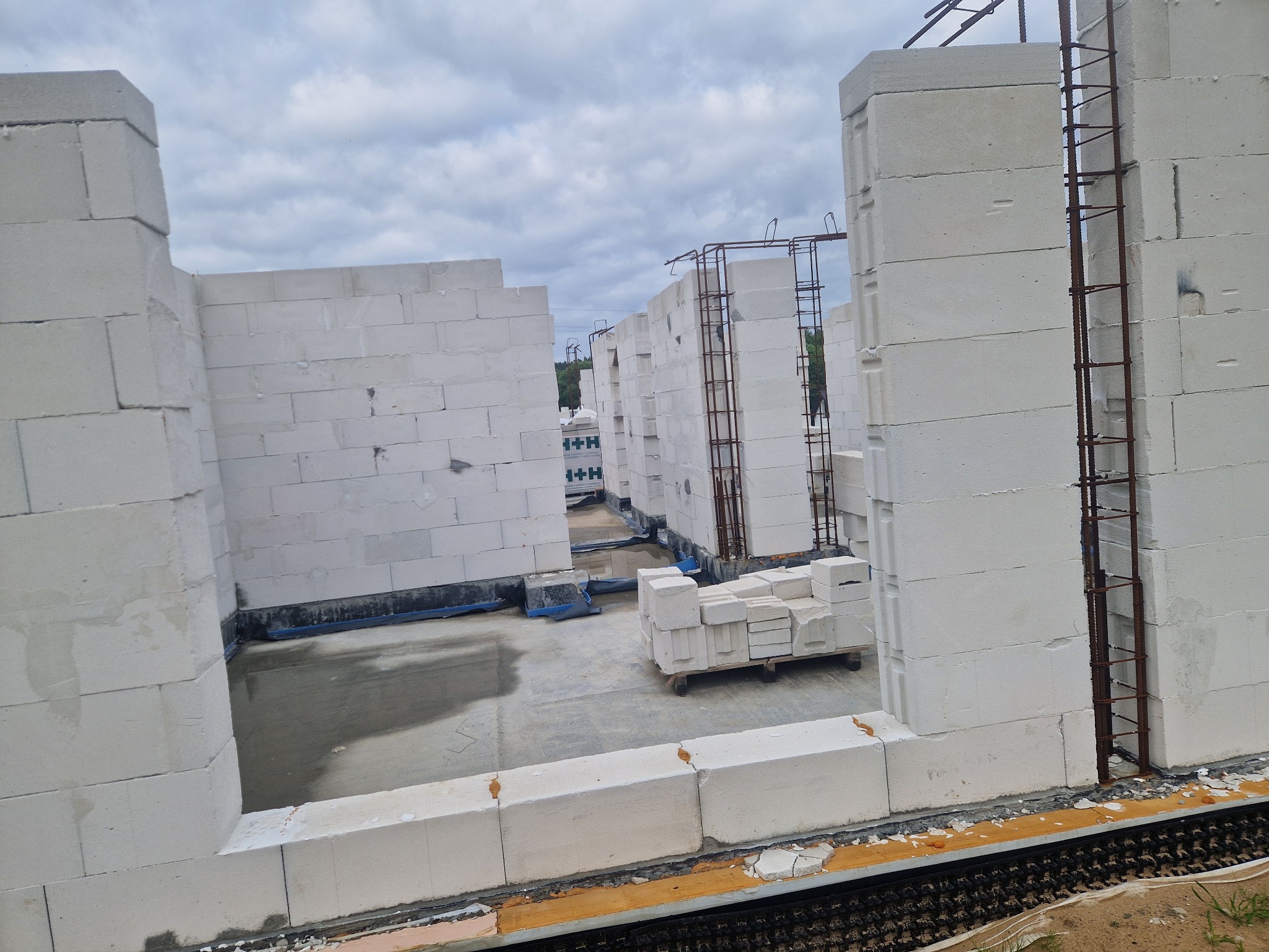 Wnętrze surowego budynku z betonu komórkowego w trakcie budowy. Widoczne ściany, zbrojenia, wózek z bloczkami i mokra posadzka.