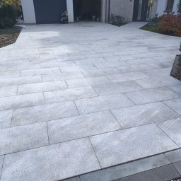 SobBruk Wiesław Sobczyk - Jasnoszara kostka granitowa ułożona na podjeździe przed nowoczesnym domem z garażem. Widoczny fragment obrzeża i osoba siedząca przed garażem.