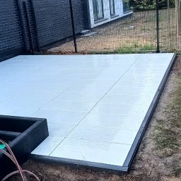 SobBruk Wiesław Sobczyk - Jasnoszara, kwadratowa nawierzchnia z płytek ułożona na zewnątrz, obok czarnej ściany budynku i ogrodzenia. Część narzędzi przykryta zieloną folią. Osoba w tle.