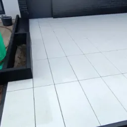 Nowoczesny taras z białych płyt, obramowany czarną listwą, przylegający do budynku z czarną cegłą. Widoczny fragment rabaty z ziemią i zielenią. Minimalistyczny design.