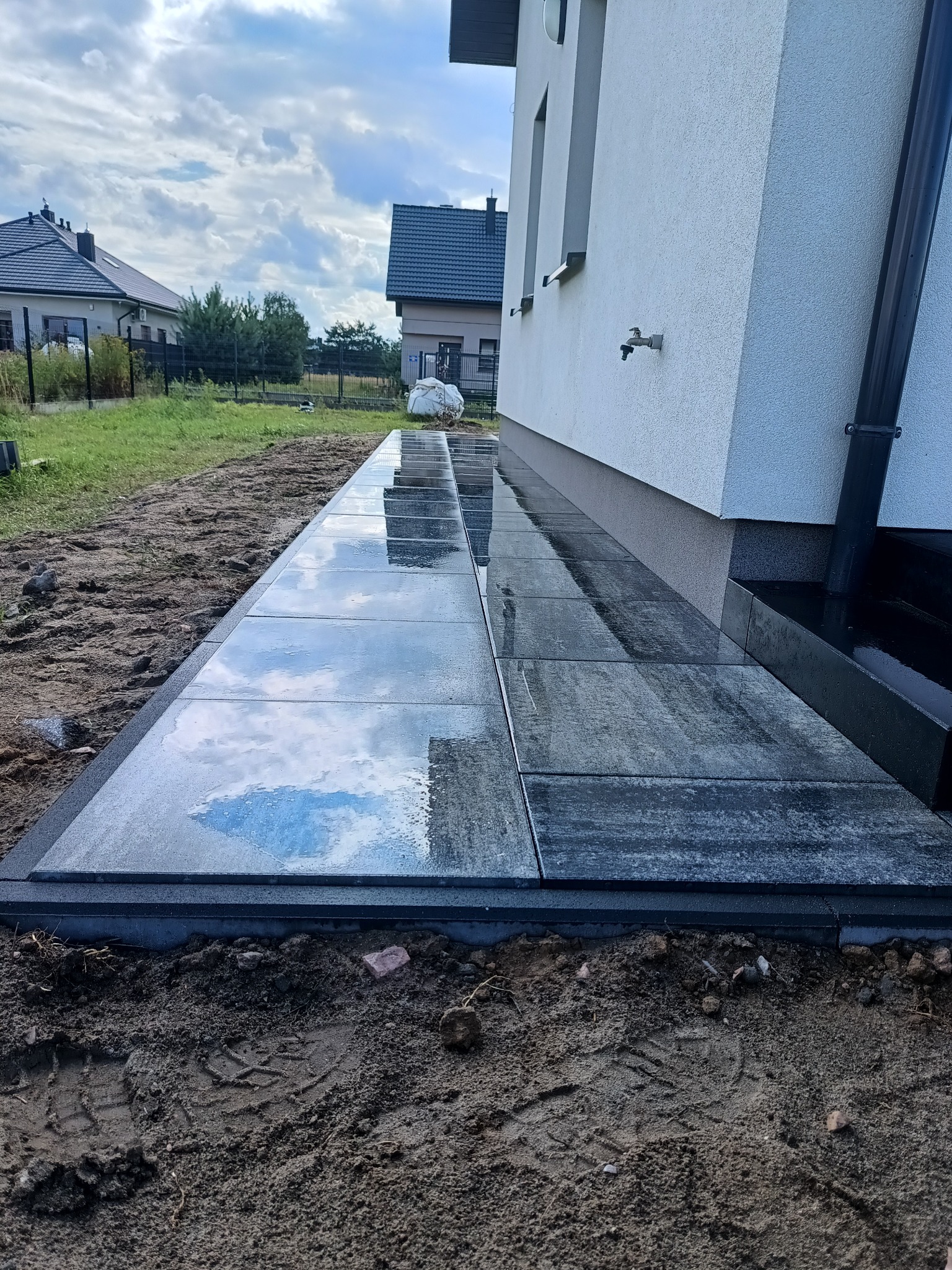 Ułożona, ciemnoszara kostka brukowa wzdłuż ściany budynku, odbijająca niebo. Widoczny fragment domu z rynną i trawnik w tle.