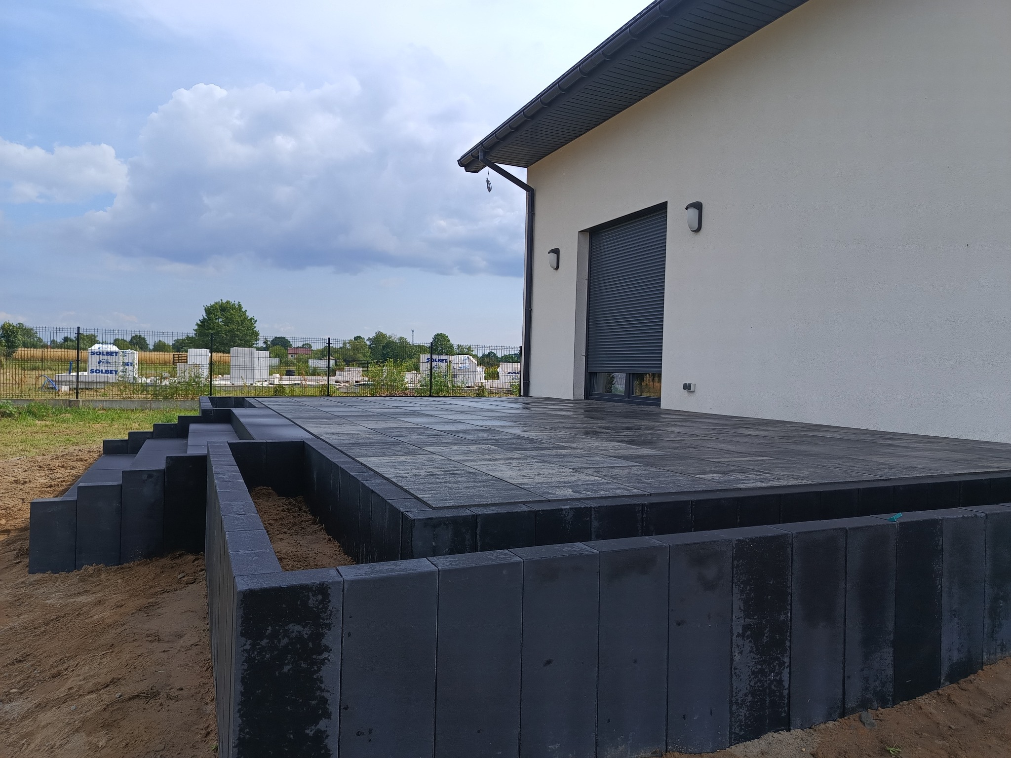 Nowoczesny taras z ciemnej kostki brukowej przylegający do białego budynku, z elementami schodów i obrzeży, na tle pochmurnego nieba i zieleni.