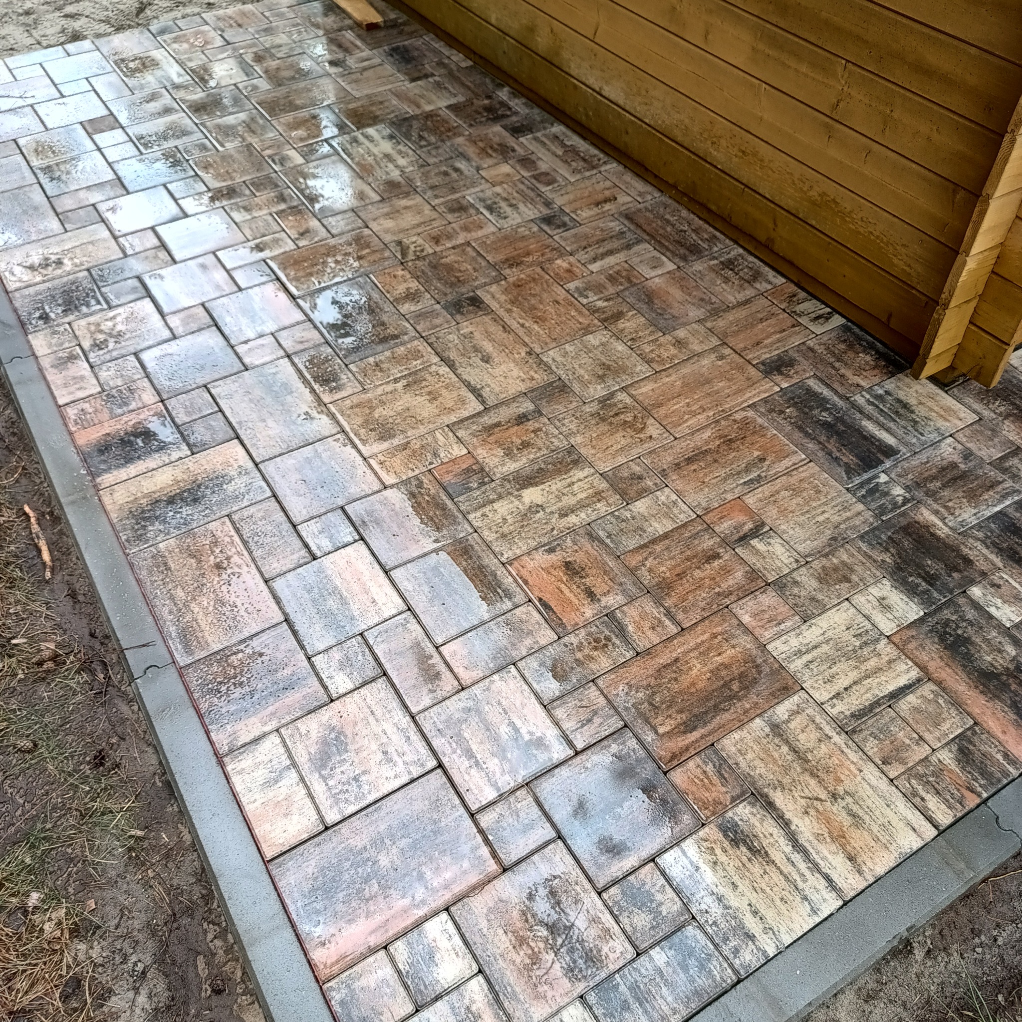 Ułożona, wielokolorowa kostka brukowa przy drewnianej konstrukcji. Widoczne szare obrzeża i mokra nawierzchnia z refleksami światła.