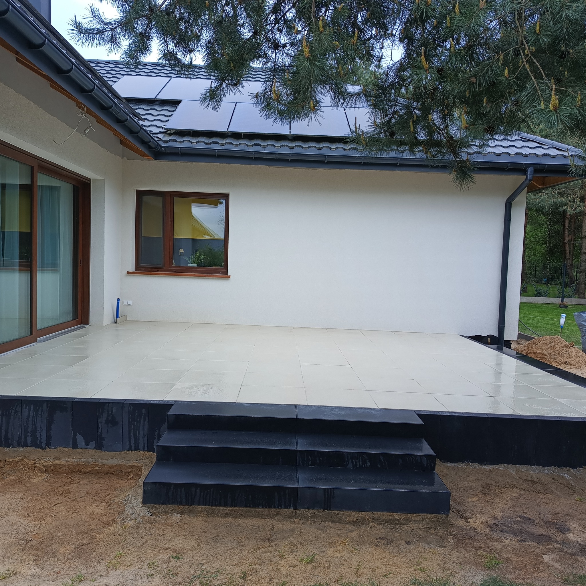 Wykończony taras z jasnych płyt, z czarnymi schodami i obrzeżem. Widoczne panele słoneczne na dachu domu i drzewo iglaste w tle. Ujęcie z poziomu gruntu.