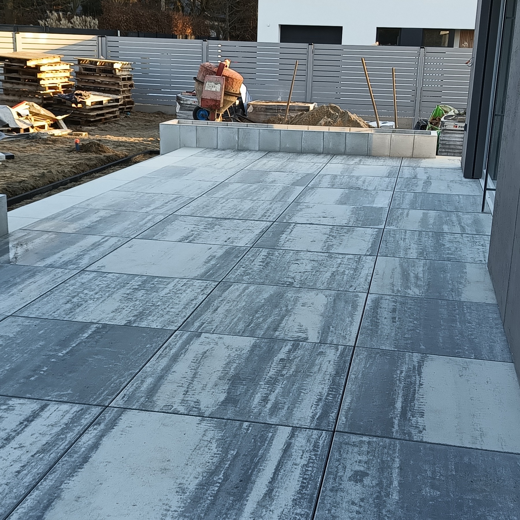 Nowoczesny taras wyłożony szarą kostką. W tle widoczny betonowy murek, mieszalnik do betonu i palety. Całość w jasnym oświetleniu.
