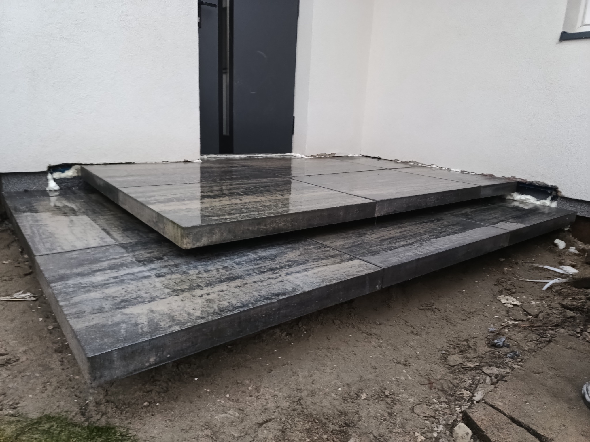 Dwu-stopniowe schody wejściowe z grafitowej kostki brukowej, wykończone pianką montażową, prowadzące do ciemnych drzwi w białym budynku. Widok z dołu.