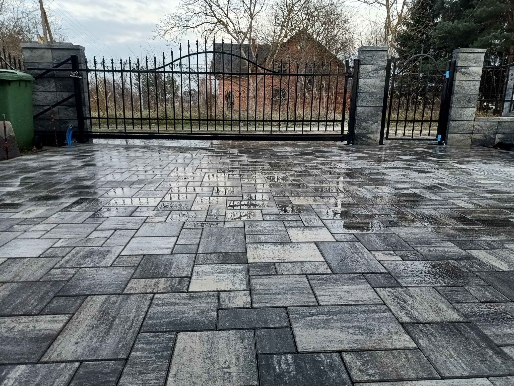 Ułożona, szara kostka brukowa na podjeździe z metalową bramą w tle. Widoczne refleksy wody na powierzchni kostki. Pochmurne niebo.
