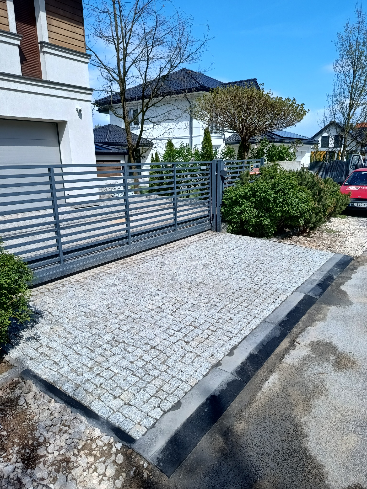 Podjazd z kostki granitowej w Nowym Dworze Mazowieckim. Szara kostka ułożona przy nowoczesnej bramie wjazdowej. Widoczne obrzeża i fragment posesji.