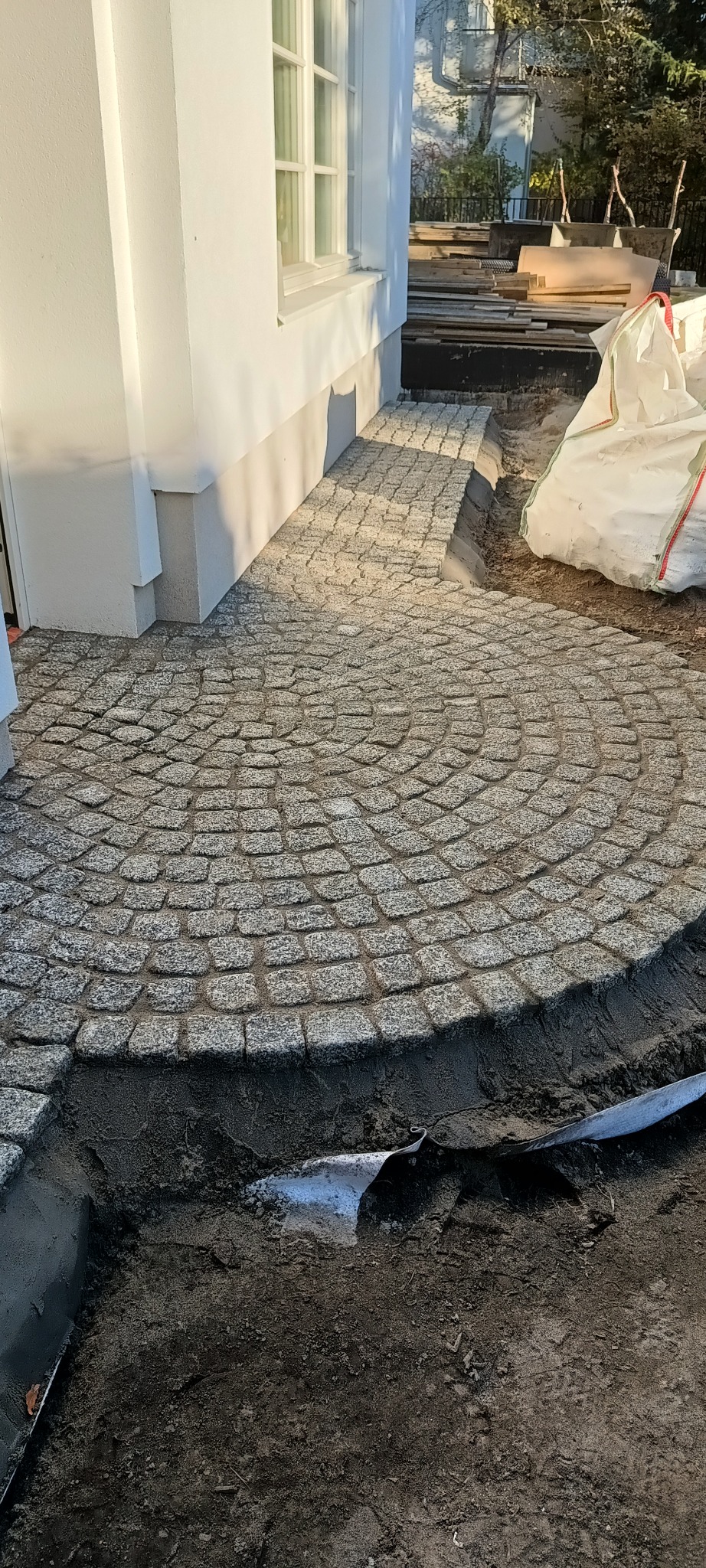 Okrągły wzór z kostki granitowej ułożony przy białym budynku. Widoczne przygotowania do dalszych prac, worki z materiałem i narzędzia w tle.