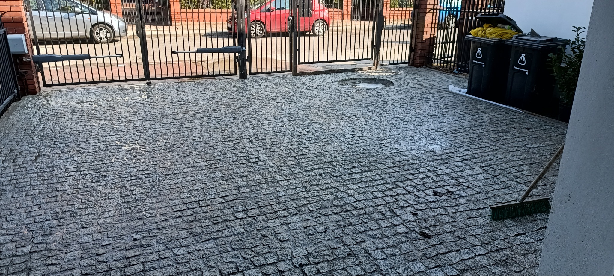 Szara kostka granitowa ułożona na podjeździe przed bramą wjazdową. Widoczne dwa kosze na śmieci i szczotka. W tle zaparkowane samochody.