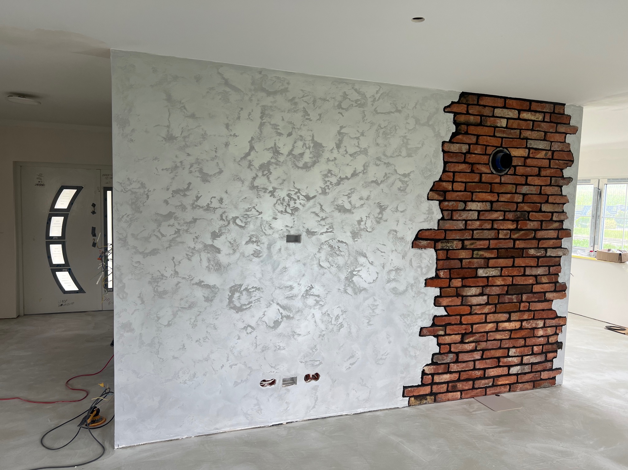 Wykończenie ściany z efektem dekoracyjnym: szpachlowana struktura w odcieniach szarości i fragment ściany z cegły z otworem wentylacyjnym. Surowy stan podłogi.