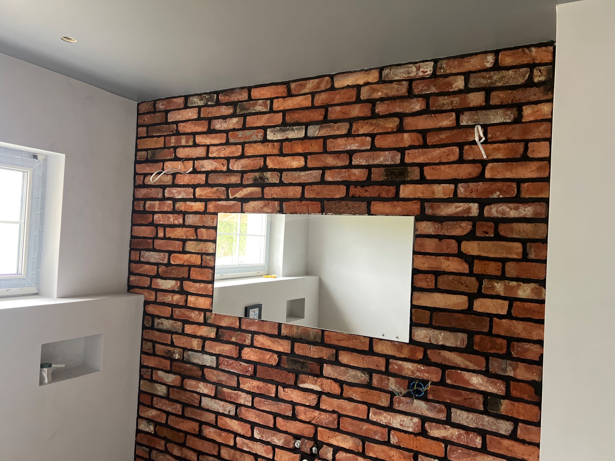 Ściana z cegły z lustrem w łazience, widoczne przewody elektryczne i fragment ściany pomalowanej na szaro. Dekoracyjne wykończenie wnętrza w stylu industrialnym.