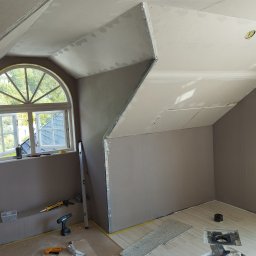 DAMIAN RYBAK - Adaptacja poddasza: montaż płyt gipsowo-kartonowych na skośnym suficie, widoczne narzędzia i okno łukowe. Etap szpachlowania łączeń płyt.