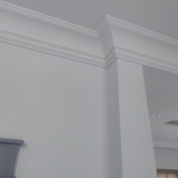 DAMIAN RYBAK - Białe, eleganckie listwy dekoracyjne na styku ściany i sufitu, tworzące estetyczne wykończenie wnętrza. Widoczny fragment ściany i kolumna.
