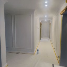 DAMIAN RYBAK - Korytarz po malowaniu na biało, z dekoracyjnymi panelami ściennymi i zabezpieczeniami podłogi taśmą malarską. Widoczne oświetlenie sufitowe i butelka wody na podłodze.