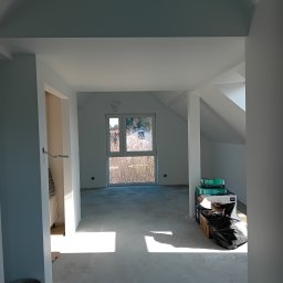 Jasne, przestronne poddasze po malowaniu na biało, z oknem i stertą materiałów budowlanych. Widoczne ślady prac wykończeniowych, surowy beton na podłodze.