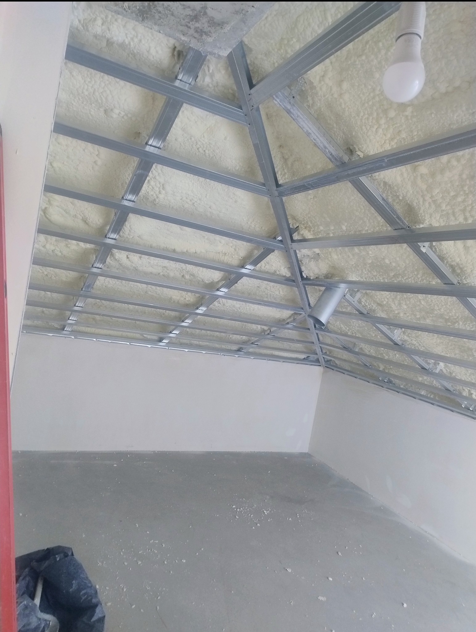 Sufit podwieszany ze stalową konstrukcją i izolacją piankową, widok z dołu. W tle surowa ściana i podłoga, widoczna żarówka. Ujęcie z dołu, perspektywa podkreśla geometrię konstrukcji.