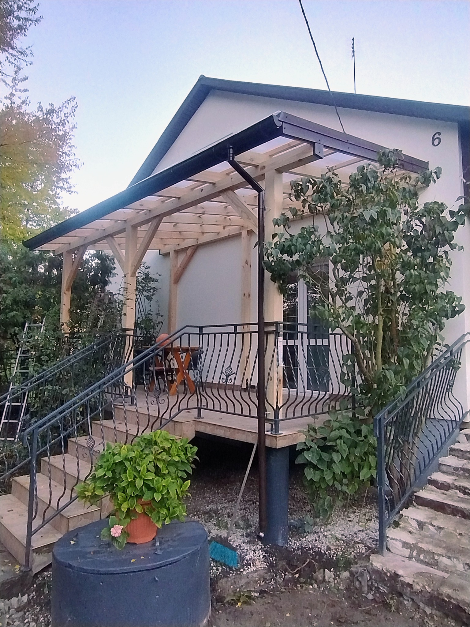 Zadaszony taras z drewnianą konstrukcją i metalową balustradą przy domu z numerem 6. Roślinność w tle i donica z kwiatami na pierwszym planie.