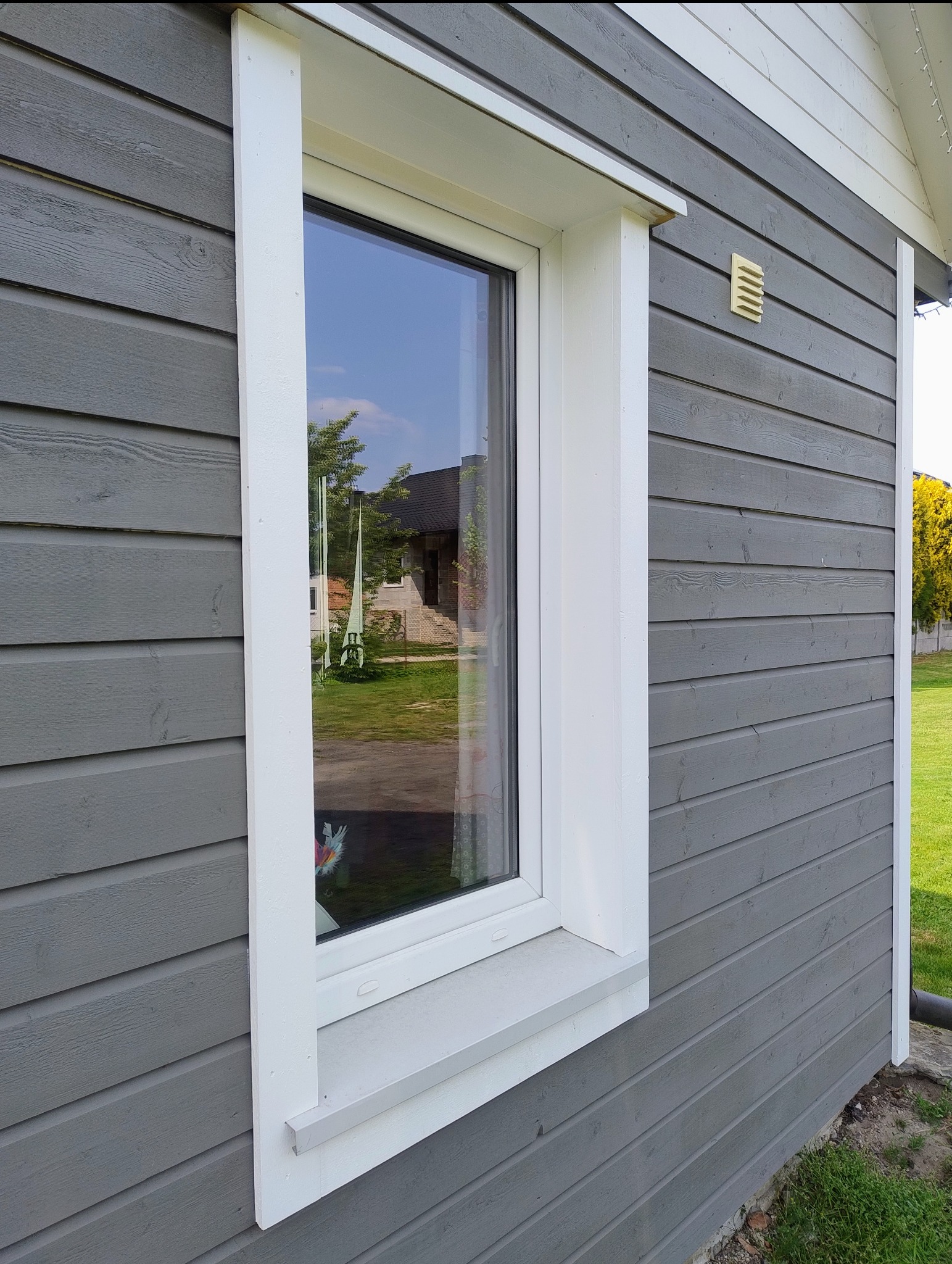 Szare drewniane deski elewacyjne z białym obramowaniem okna. W oknie odbija się budynek i zieleń. Widoczny fragment rynny i kratka wentylacyjna.