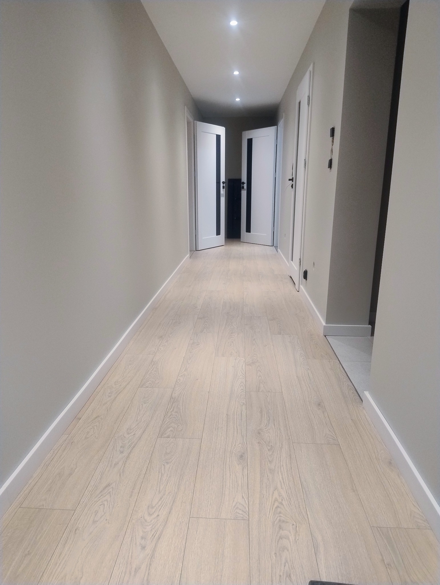 Jasny korytarz z panelami podłogowymi i białymi listwami przypodłogowymi. W tle otwarte białe drzwi i oświetlenie sufitowe.