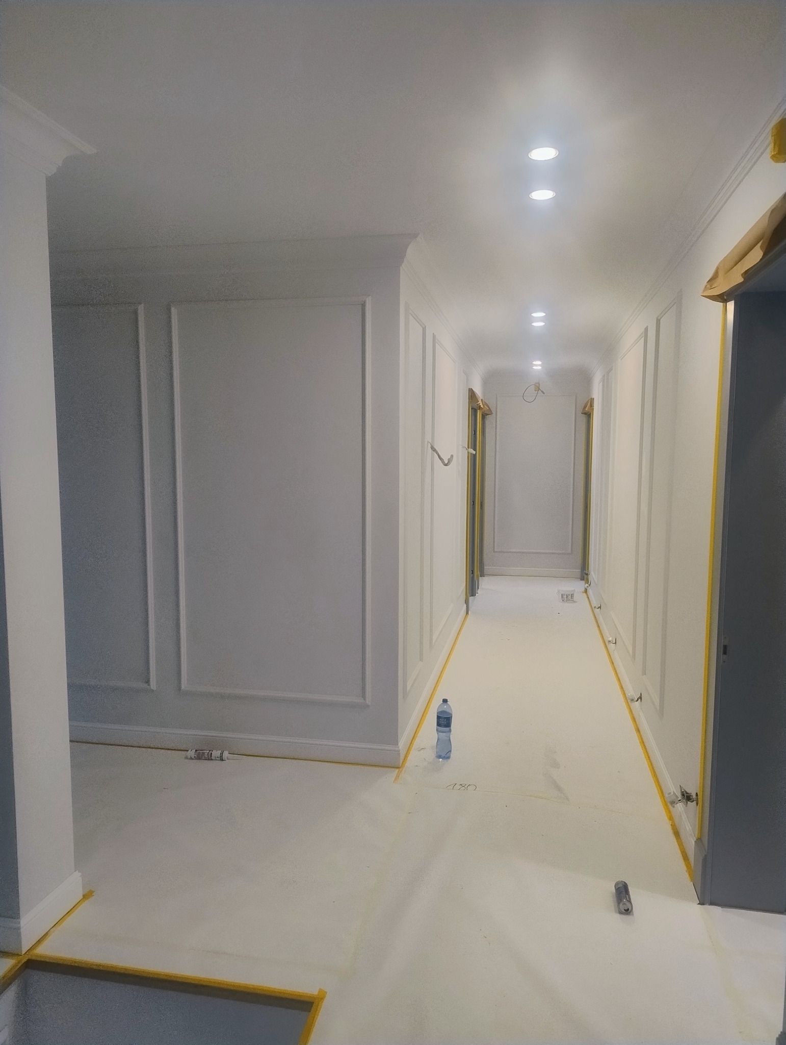 Korytarz po malowaniu na biało, z dekoracyjnymi panelami ściennymi i zabezpieczeniami podłogi taśmą malarską. Widoczne oświetlenie sufitowe i butelka wody na podłodze.