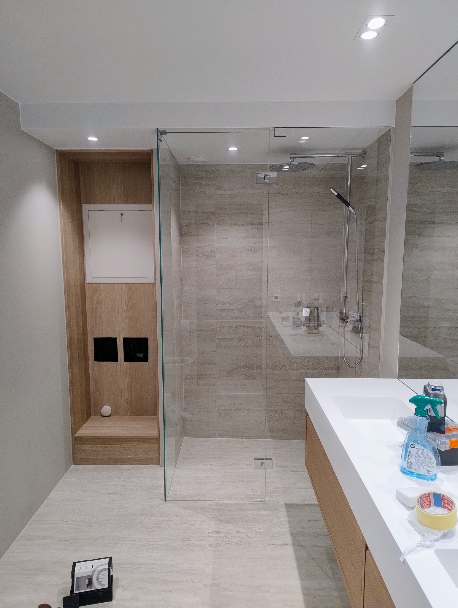 Nowoczesna łazienka z kabiną prysznicową typu walk-in, beżowe płytki imitujące kamień, zabudowa z drewnianymi akcentami i białą umywalką. Minimalistyczny design.