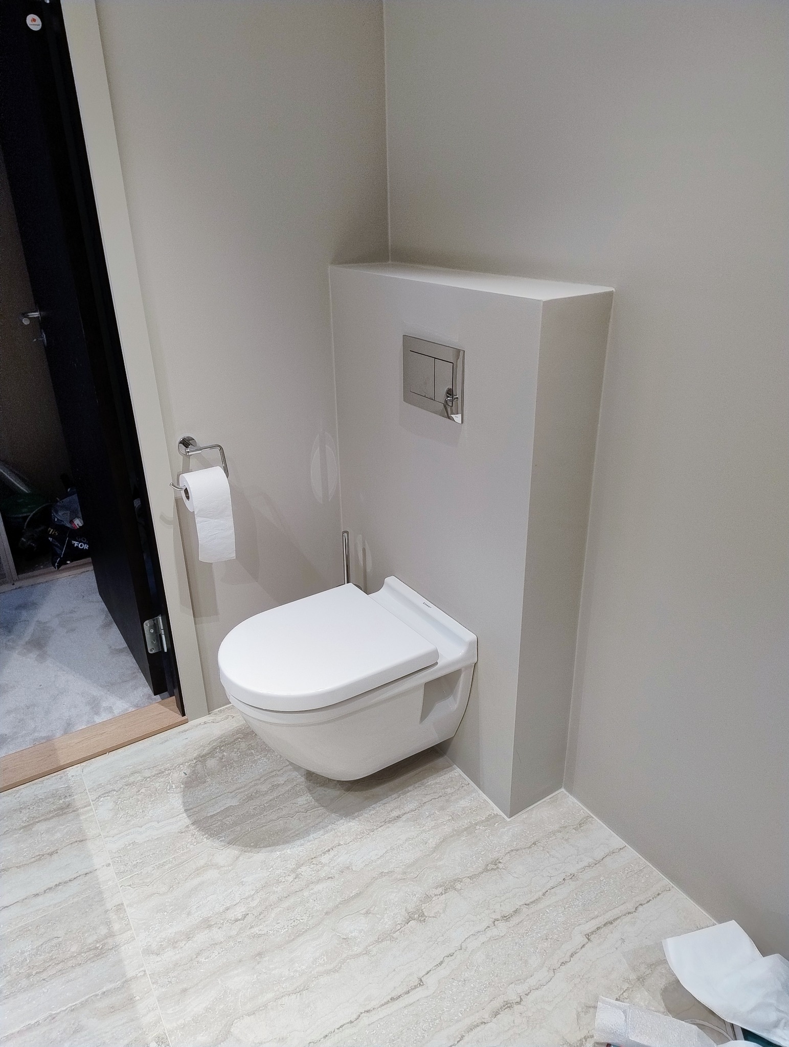 Nowoczesna, podwieszana toaleta w jasnej łazience z beżowymi płytkami imitującymi marmur. Widoczny uchwyt na papier toaletowy i zabudowany stelaż.