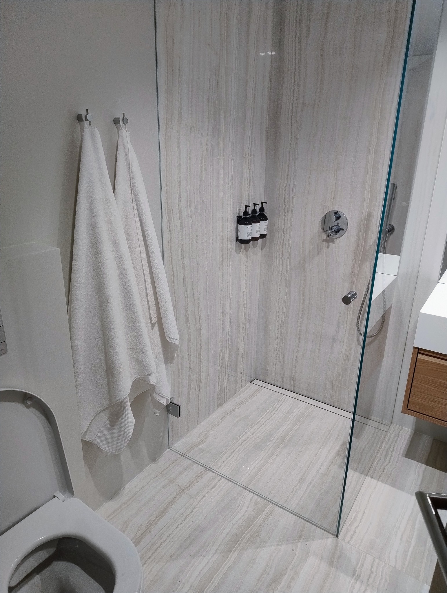 Nowoczesna łazienka z kabiną prysznicową typu walk-in, wykończona płytkami imitującymi marmur. Widoczne białe ręczniki na wieszakach i minimalistyczna armatura.