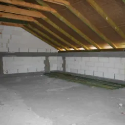 Surowe poddasze z widoczną konstrukcją dachu, ścianami z betonu komórkowego i stertą zielonych łat. Wnętrze w trakcie budowy lub remontu.