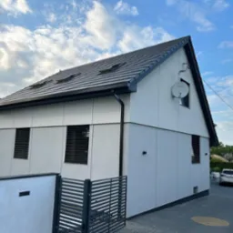 Nowoczesny dom jednorodzinny z szarą elewacją i ciemną dachówką, widok z zewnątrz na tle błękitnego nieba z chmurami. Widoczne detale: antena satelitarna, żaluzje.